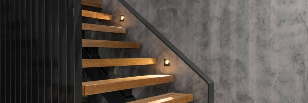 Your Guide to Cement Render: Everything You Need to Know black staircase with wooden stairs and warm lights on a grey textured wall